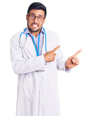 Wall Mural - Young hispanic man wearing doctor uniform and stethoscope pointing aside worried and nervous with both hands, concerned and surprised expression