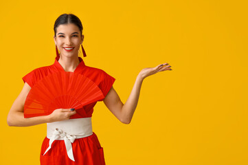 Poster - Beautiful young happy woman with fan pointing at something on yellow background. Chinese New Year celebration