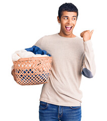 Canvas Print - Young african amercian man holding laundry basket pointing thumb up to the side smiling happy with open mouth