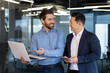 © Liubomir - Two businessmen men of different races are standing in the office, looking at a laptop, discussing work topics, smiling at each other.
