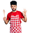 © Krakenimages.com - Young arab man with beard wearing baker uniform holding wooden spoon in shock face, looking skeptical and sarcastic, surprised with open mouth
