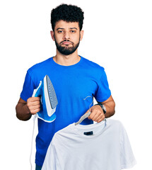 Wall Mural - Young arab man with beard holding electric steam iron and white t shirt depressed and worry for distress, crying angry and afraid. sad expression.