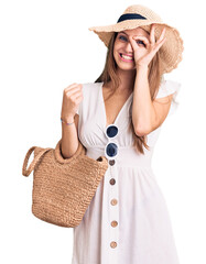 Young beautiful blonde woman wearing summer dress and wicker handbag smiling happy doing ok sign with hand on eye looking through fingers