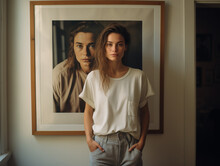A Young Woman Wearing White T-shirt And Grey Trousers, Is Standing In Front Of A Bit Portrait Of Herself, Hanging On The Wall. Beauty And Modelling Concept. Generative AI