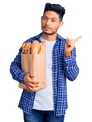 Wall Mural - Handsome latin american young man holding paper bag with bread pointing with hand finger to the side showing advertisement, serious and calm face