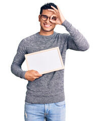 Wall Mural - Hispanic handsome young man holding blank empty banner smiling happy doing ok sign with hand on eye looking through fingers