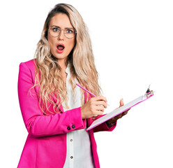 Canvas Print - Young blonde woman writing on clipboard in shock face, looking skeptical and sarcastic, surprised with open mouth