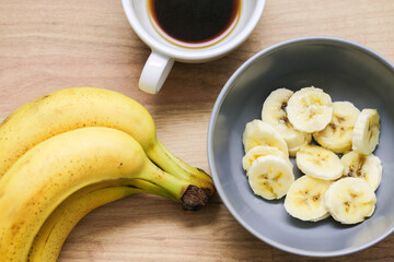 Top view banana salad with coffee. Bowl with fruit slices inside. Banana slices in bowl. Wooden table healthy breakfast. Banana pieces background. White ceramic coffee cup. Morning breakfast.