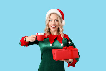 Sticker - Beautiful young woman dressed as elf holding Christmas gift and cup of hot chocolate with marshmallows on blue background