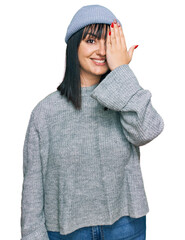 Wall Mural - Young hispanic woman wearing cute wool cap covering one eye with hand, confident smile on face and surprise emotion.