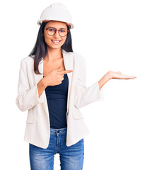 Poster - Young beautiful latin girl wearing architect hardhat and glasses amazed and smiling to the camera while presenting with hand and pointing with finger.