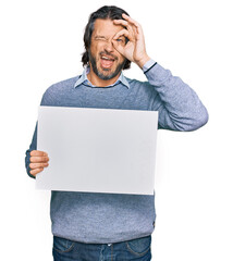 Poster - Middle age handsome man holding blank empty banner smiling happy doing ok sign with hand on eye looking through fingers