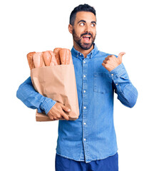 Poster - Young hispanic man holding bag with bread pointing thumb up to the side smiling happy with open mouth