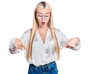 Beautiful blonde woman wearing elegant shirt and glasses pointing down with fingers showing advertisement, surprised face and open mouth