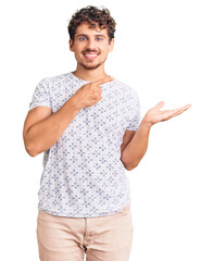 Wall Mural - Young handsome man with curly hair wearing casual clothes amazed and smiling to the camera while presenting with hand and pointing with finger.