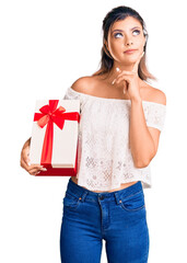 Poster - Young beautiful woman holding gift serious face thinking about question with hand on chin, thoughtful about confusing idea