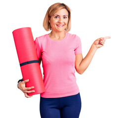 Poster - Young blonde woman holding yoga mat smiling happy pointing with hand and finger to the side