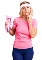 Wall Mural - Young blonde woman wearing sportswear holding water bottle serious face thinking about question with hand on chin, thoughtful about confusing idea