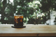 Cold black coffee with ice cubes on a wood table against nature background. Cold Brew. Summer drinks.
