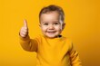 © Iftikhar alam - Portrait of a smiling baby boy showing thumbs up over yellow background, a toddler giving a thumbs up on yellow background, AI Generated