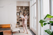 © Iona - Full body image of young brunette businesswoman in suit standing at home holds paper sheets, exploring documents standing barefoot at luxury apartment with book shelf and fireplace.