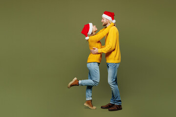 Full body cheerful lovely merry young couple two friends man woman wears sweater Santa hat posing look camera hug cuddle isolated on plain green background. Happy New Year Christmas holiday concept.