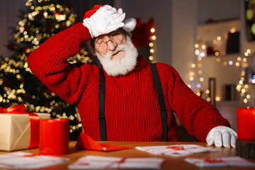 Poster - Tired Santa Claus at his workplace. Letters and gift box on table in room with Christmas tree