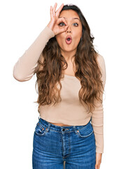 Wall Mural - Young hispanic girl wearing casual clothes doing ok gesture shocked with surprised face, eye looking through fingers. unbelieving expression.