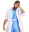 Young hispanic man wearing doctor uniform and stethoscope doing ok gesture shocked with surprised face, eye looking through fingers. unbelieving expression.