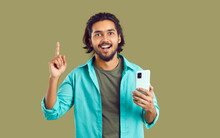 Young Attractive Indian Man Smiling Rejoicing At New Idea And Holding Phone To Make Note Of Upcoming Plans Showing Finger Up Dressed In Casual Style Stands On Olive Background. Concept Planning App