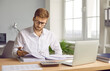© Studio Romantic - Portrait of young serious businessman in glasses working at home or at office. Man looking through paper financial documents with a pile of folders on his workplace analyzing company.