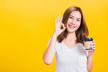 Wall Mural - Portrait Asian Thai beautiful happy young woman standing holds take away cup coffee paper and confident show OK sign with fingers excellent, studio shot isolated on yellow background, with copy space