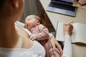 top down view at sleeping newborn with pacifier in arms of mother working and writing down ideas, co
