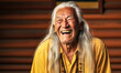 Joyful elderly Native American man with long white hair, laughing heartily, wearing a yellow shirt against a yellow wooden background, exuding warmth and vitality