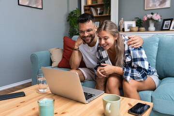 Happy young couple at home man surprise woman to choose travel destination, searching the internet on laptop computer. Online booking. Anniversary vacation and celebration of togetherness and love