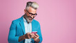 © Mangevigne - Elegant man wearing a blue jacket and glasses with tattoo and beard looking at his mobile phone and smiling on a plain pink background