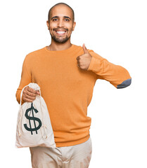 Wall Mural - Hispanic adult man holding dollars bag smiling happy and positive, thumb up doing excellent and approval sign