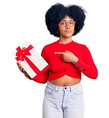 Poster - Young african american girl holding gift pointing with hand finger to the side showing advertisement, serious and calm face