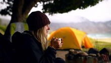 Campfire, relax and woman with coffee at tent for adventure in nature at ocean, mountain and trees. Camping, morning drink at fire and girl on travel holiday at beach with freedom, peace and thinking
