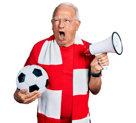 Canvas Print - Senior man with grey hair football hooligan holding ball and using megaphone angry and mad screaming frustrated and furious, shouting with anger. rage and aggressive concept.