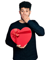 Canvas Print - Young arab man with beard holding valentine gift covering mouth with hand, shocked and afraid for mistake. surprised expression