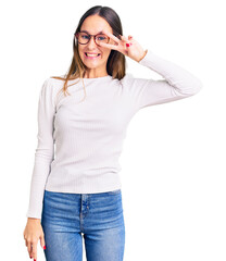 Wall Mural - Beautiful brunette young woman wearing casual white sweater and glasses doing peace symbol with fingers over face, smiling cheerful showing victory