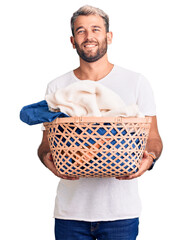 Poster - Young handsome blond man holding laundry basket with clothes looking positive and happy standing and smiling with a confident smile showing teeth