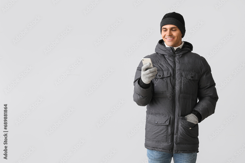 Young man in puffer jacket using mobile phone on light background