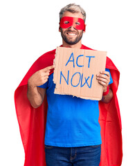 Poster - Young blond man wearing super hero costume holding act now cardboard banner smiling happy pointing with hand and finger