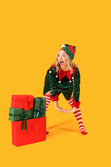 Wall Mural - Shocked young woman dressed as elf with Christmas gifts on yellow background