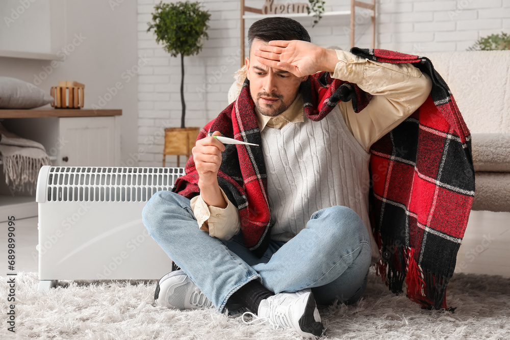 Sick young man with plaid and thermometer warming near radiator at home