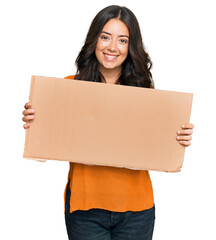 Wall Mural - Beautiful brunette young woman holding cardboard blank empty banner looking positive and happy standing and smiling with a confident smile showing teeth