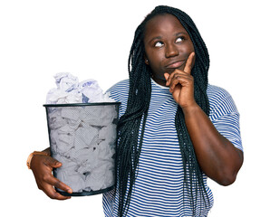 Sticker - Young black woman with braids holding paper bin full of crumpled papers serious face thinking about question with hand on chin, thoughtful about confusing idea