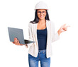 © Krakenimages.com - Young beautiful latin girl wearing architect hardhat holding laptop smiling happy pointing with hand and finger to the side